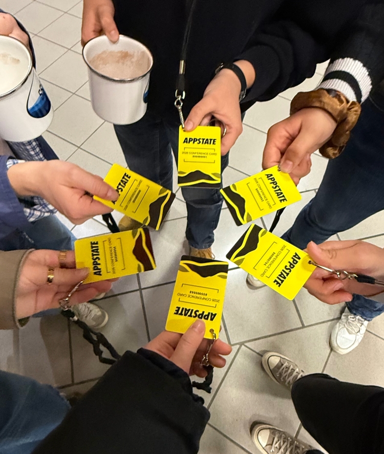 A top-down view of several hands holding yellow "APPSTATE" conference badges in a circle.