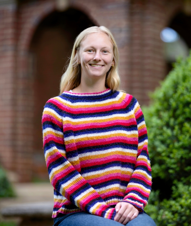 An outdoor portrait of a smiling blonde woman sitting in front of a green bush, wearing a vibrant sweater with horizontal stripes of pink, orange, white, and dark blue, with a blurred brick structure in the background.