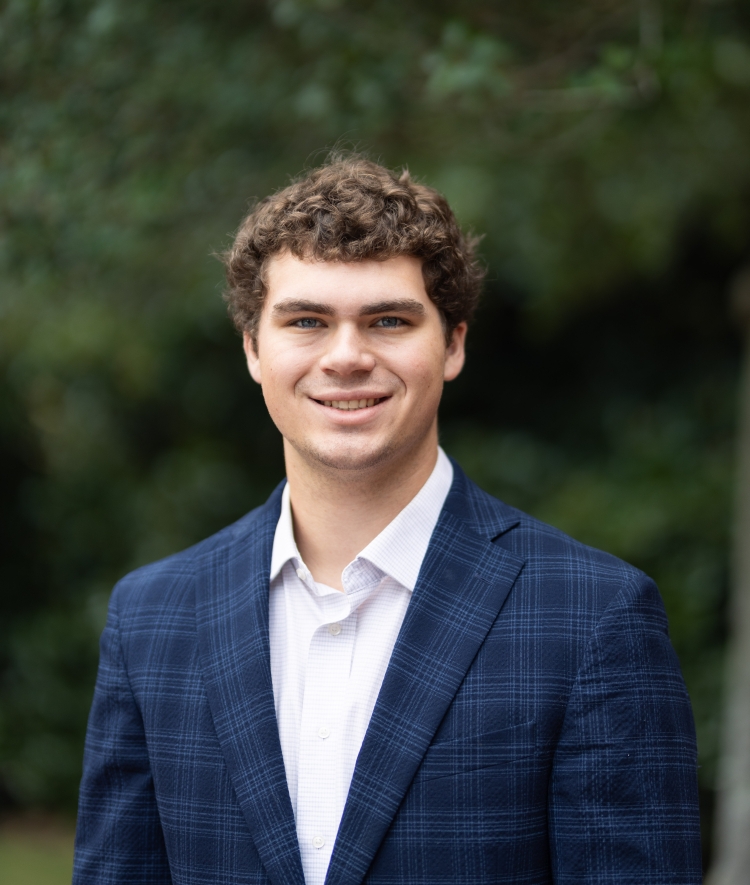 An outdoor portrait of a smiling young man with curly brown hair, wearing a blue plaid blazer and a white button-down shirt.