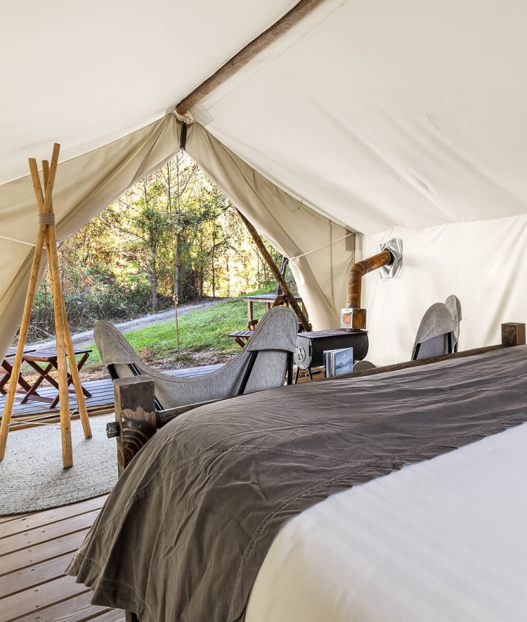 Inside a glamping tent, the view from the foot of a large bed looks out through the open canvas entryway toward two butterfly chairs on a wooden deck and a sunlit forest beyond.