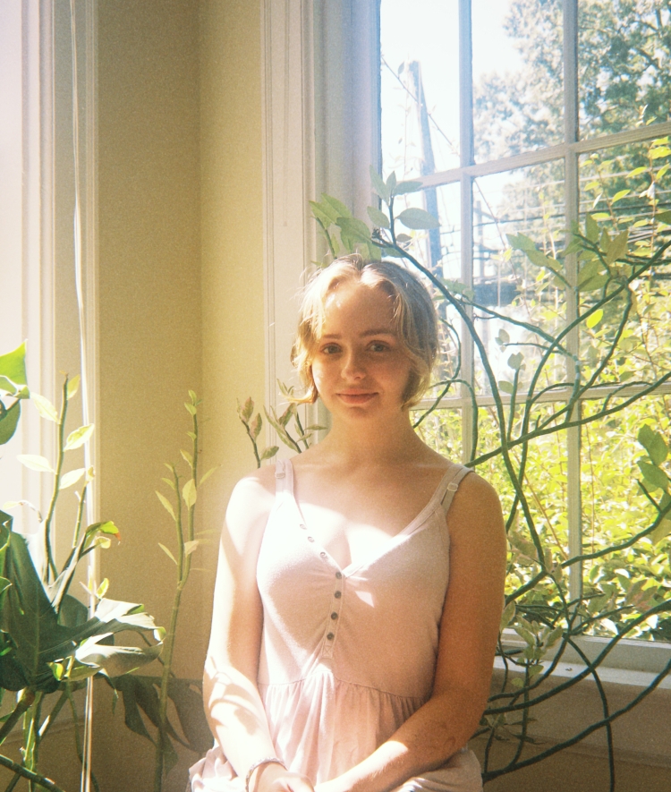 portrait of a young woman with blonde hair sitting by a bright window, surrounded by green houseplants as warm sunlight filters into the room.