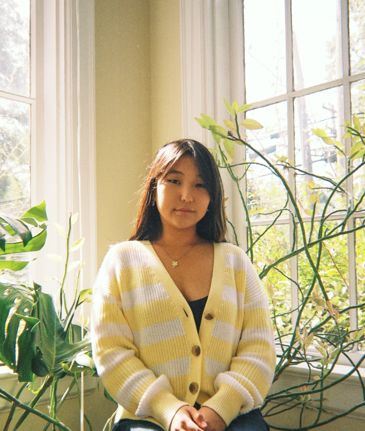 A young woman wearing a yellow and white striped cardigan sits in front of a sunny window framed by lush green plants.
