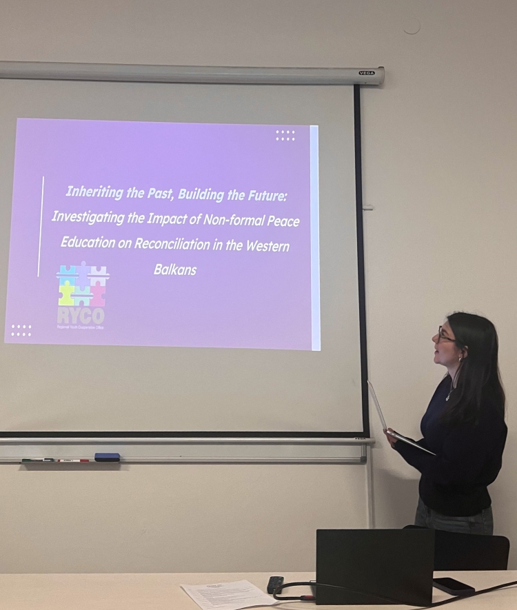 A presenter stands next to a projection screen displaying a purple presentation slide titled 'Inheriting the Past, Building the Future: Investigating the Impact of Non-formal Peace Education on Reconciliation in the Western Balkans' with the RYCO logo.