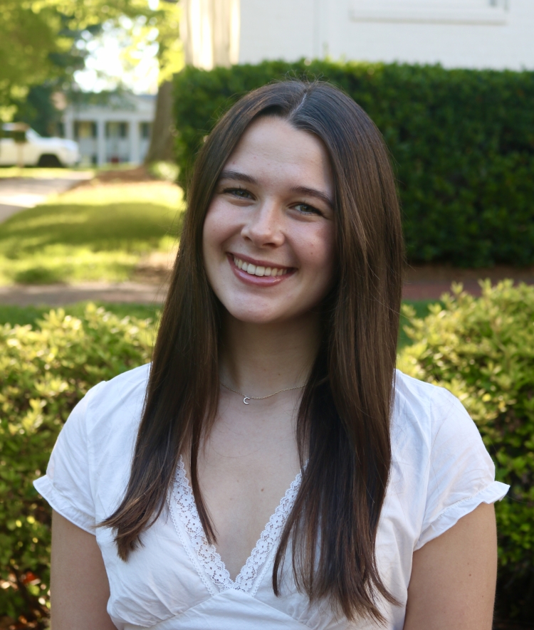 A smiling woman with long, straight dark brown hair wears a white V-neck top with lace trim, posed outdoors in front of green bushes.