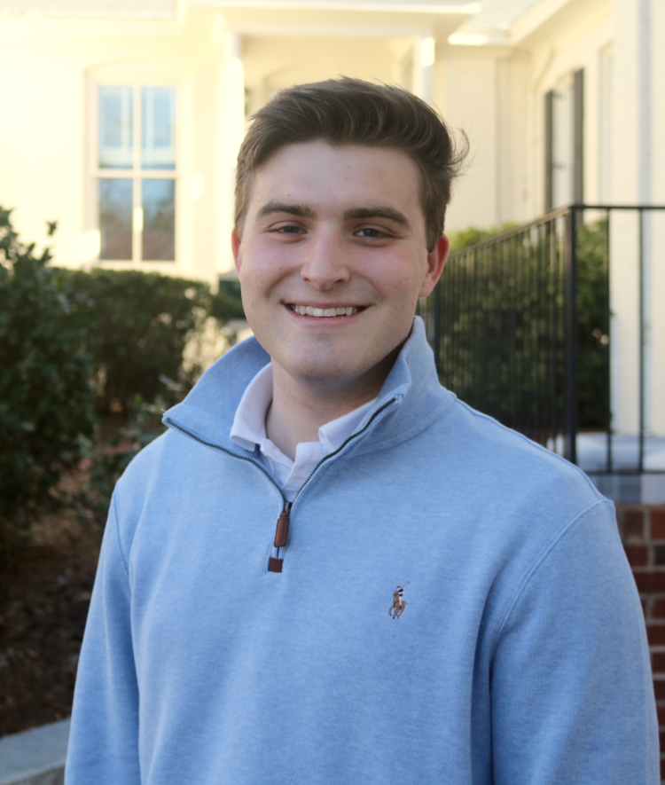 A smiling young man with styled brown hair wears a light blue quarter-zip pullover over a white collared shirt, standing outdoors in front of a blurred light-colored building.