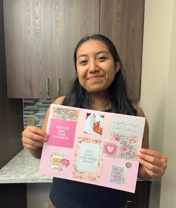 A smiling young woman holds up a pink vision board covered in motivational quotes, affirmations, and small photos against a kitchen backdrop.