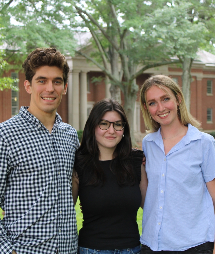 hree young adults smile and pose together arm-in-arm on a green grassy lawn with a brick college campus building and trees in the background