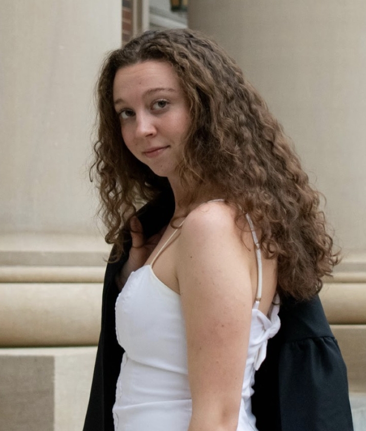 A young woman with long curly brown hair, white sundress and graduation robe