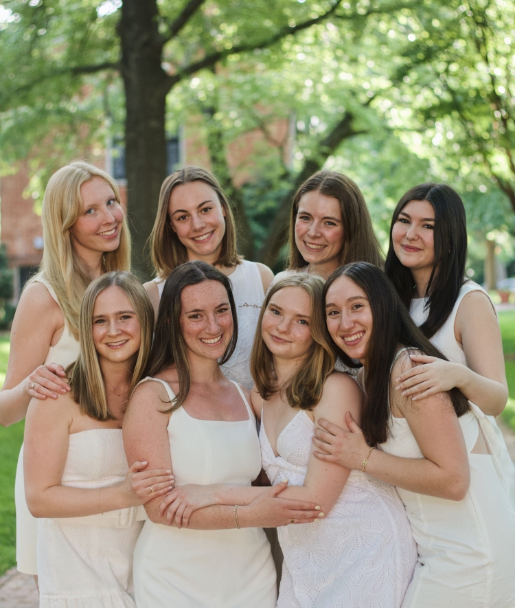 A group of eight young women in white dresses smile and embrace while posing closely together outdoors under shaded green trees