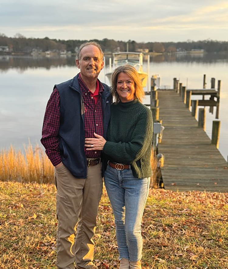 A smiling man and woman pose together on a grassy shoreline at sunset, with a wooden boat dock, a calm body of water, and a distant treeline behind them.