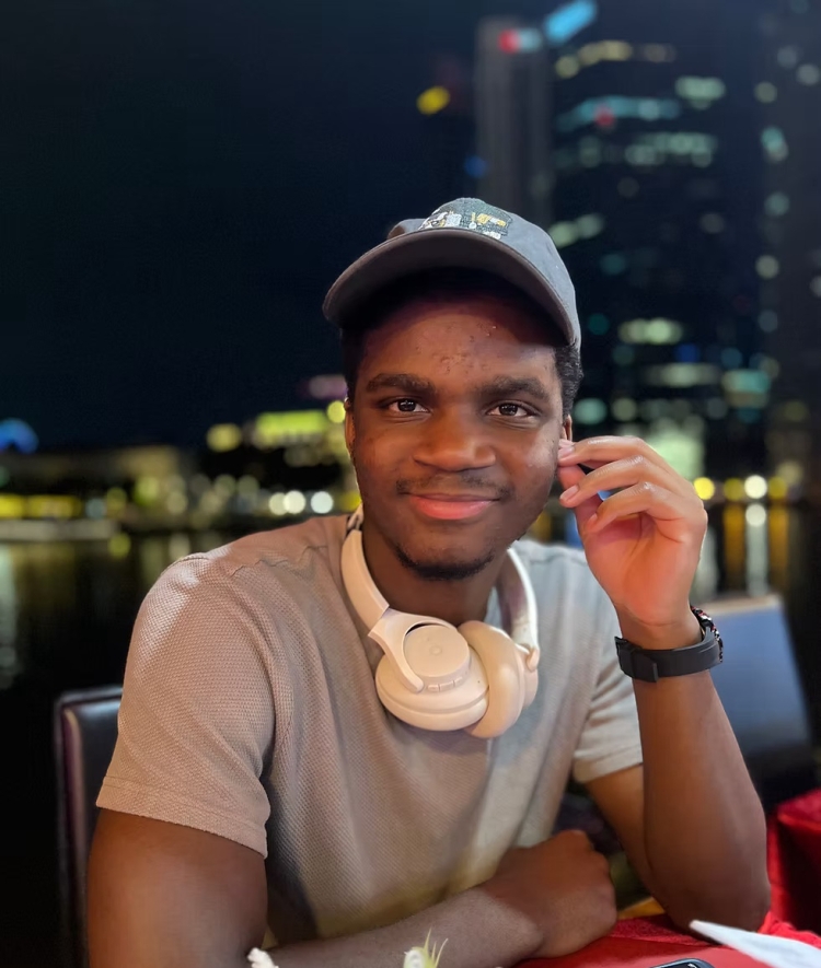 A young man wearing a baseball cap and headphones around his neck smiles while seated at an outdoor waterfront restaurant at night.