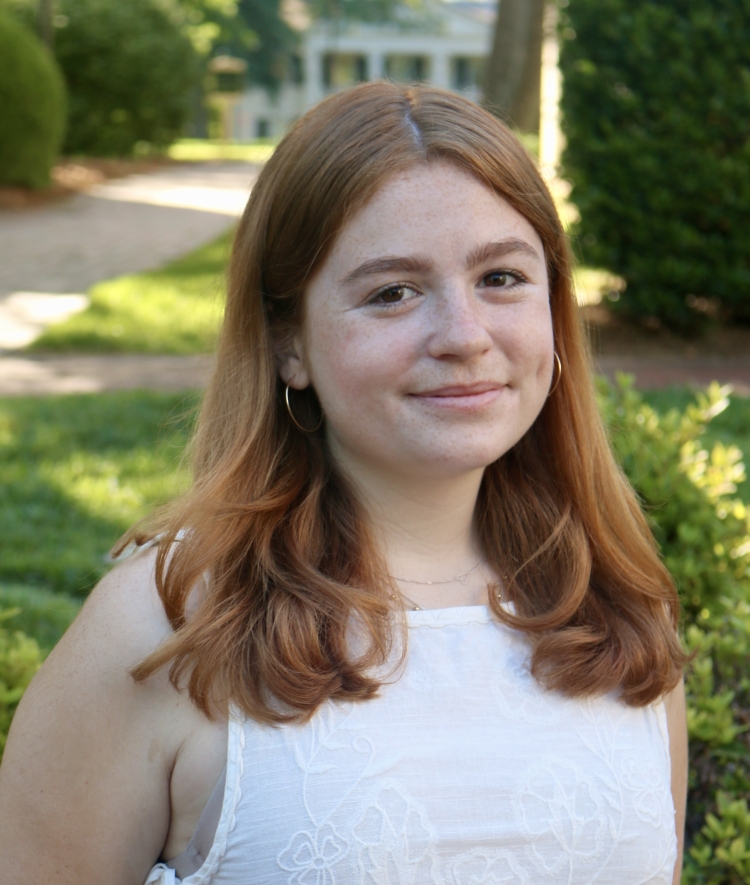 A smiling woman with shoulder-length strawberry blonde hair and freckles wears a white textured sleeveless top and small hoop earrings in an outdoor setting.