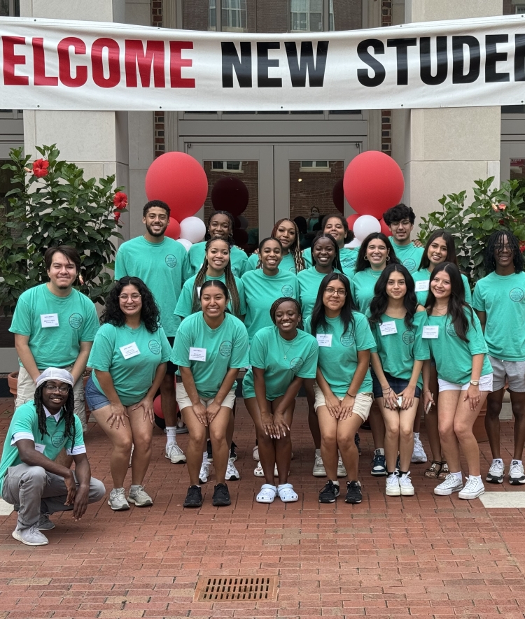 A diverse group of student leaders wearing matching teal t-shirts smiles and poses together on a brick courtyard underneath a large banner reading "WELCOME NEW STUDENTS."