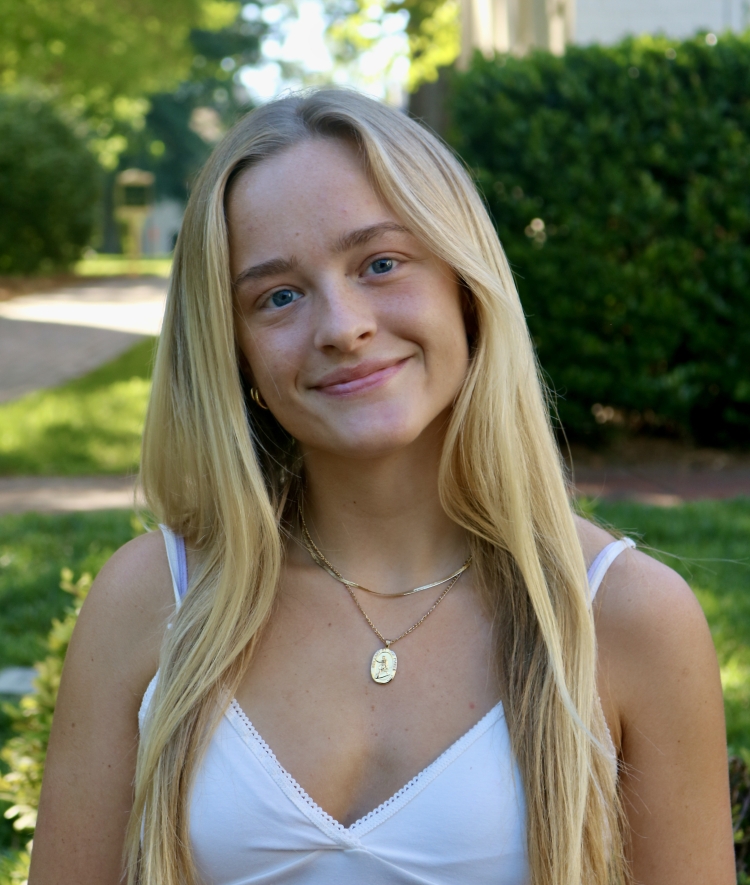 A smiling woman with long straight blonde hair and a gold necklace wears a white camisole top against a sunny, blurred outdoor backdrop.