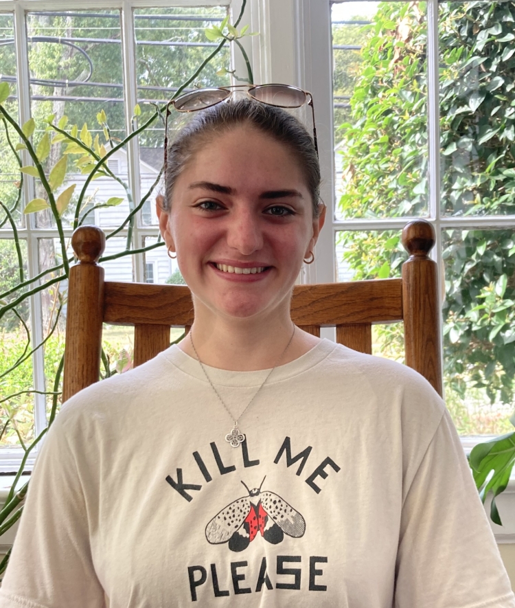 A young woman smiles while sitting in a wooden chair indoors, wearing sunglasses on her head and a t-shirt featuring a spotted lanternfly graphic