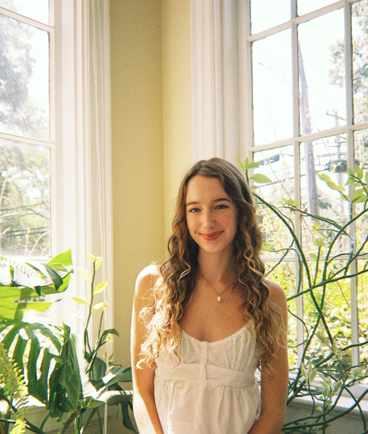 A young woman with long, curly blonde hair smiles gently while posing near a bright window flanked by large houseplants.