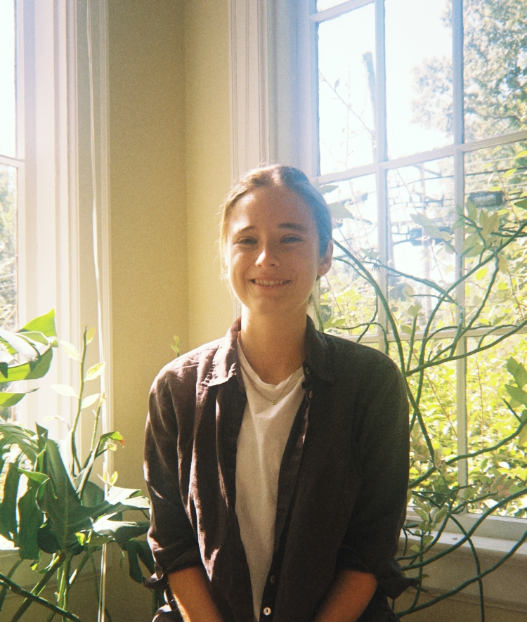 A smiling young woman with her hair pulled back sits in dappled sunlight between large windows and vibrant green plants.