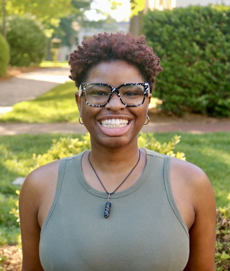 A widely smiling woman with short, curly reddish-brown hair and patterned glasses wears an olive green ribbed tank top and a crystal pendant necklace outdoors.