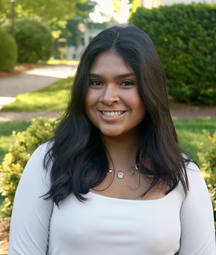 A smiling woman with long, wavy dark hair and a charm necklace wears a cream-colored square-neck long-sleeve shirt in front of outdoor greenery.