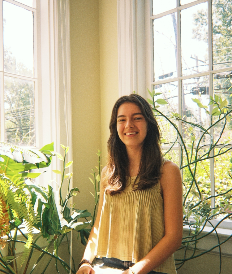 A young woman with long brown hair smiles warmly while sitting by an indoor window surrounded by leafy green plants.