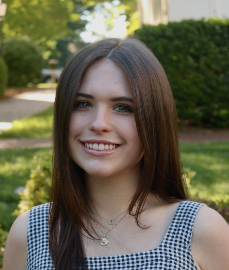 A smiling woman with long, straight dark brown hair and layered gold necklaces wears a black and white checkered sleeveless top outdoors.
