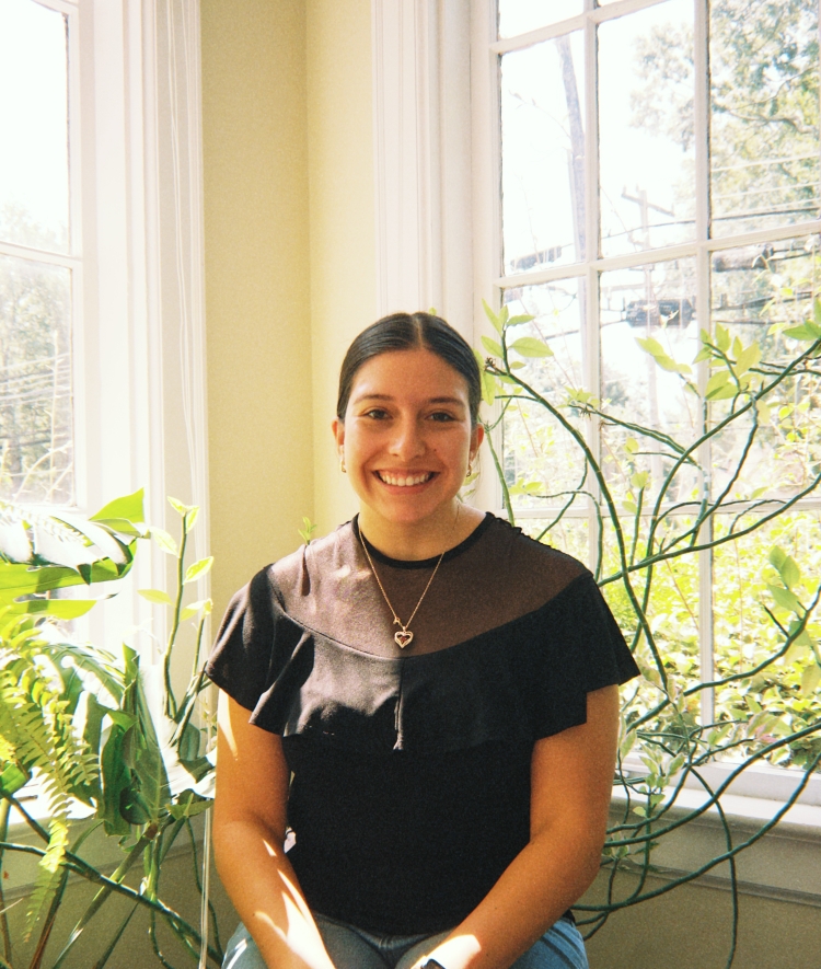 A smiling young woman in a black top with sheer details sits in front of bright windows with potted houseplants beside her.