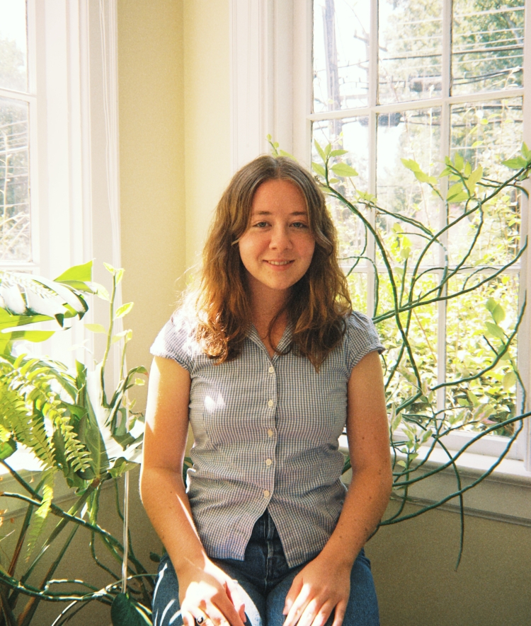 A young woman with wavy brown hair wearing a blue gingham shirt sits in bright sunlight between windows and green plants.