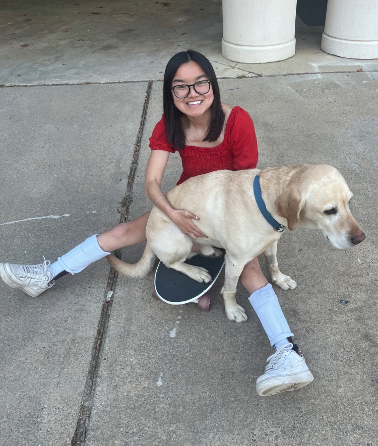 A smiling young woman in a red shirt sits on a concrete ground with her legs extended, holding a yellow Labrador Retriever that is sitting on a skateboard between her legs