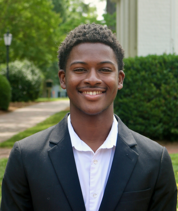 A young Black man with short, textured hair smiles warmly while wearing a black blazer and an unbuttoned white collared shirt, pictured outdoors against a blurred background of green trees and a pathway.