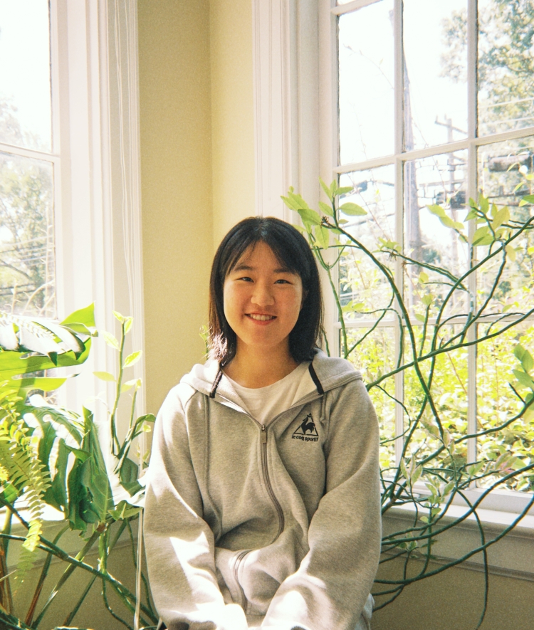 A young woman in a grey zip-up hoodie smiles while sitting indoors by a bright window with large potted plants on the side.