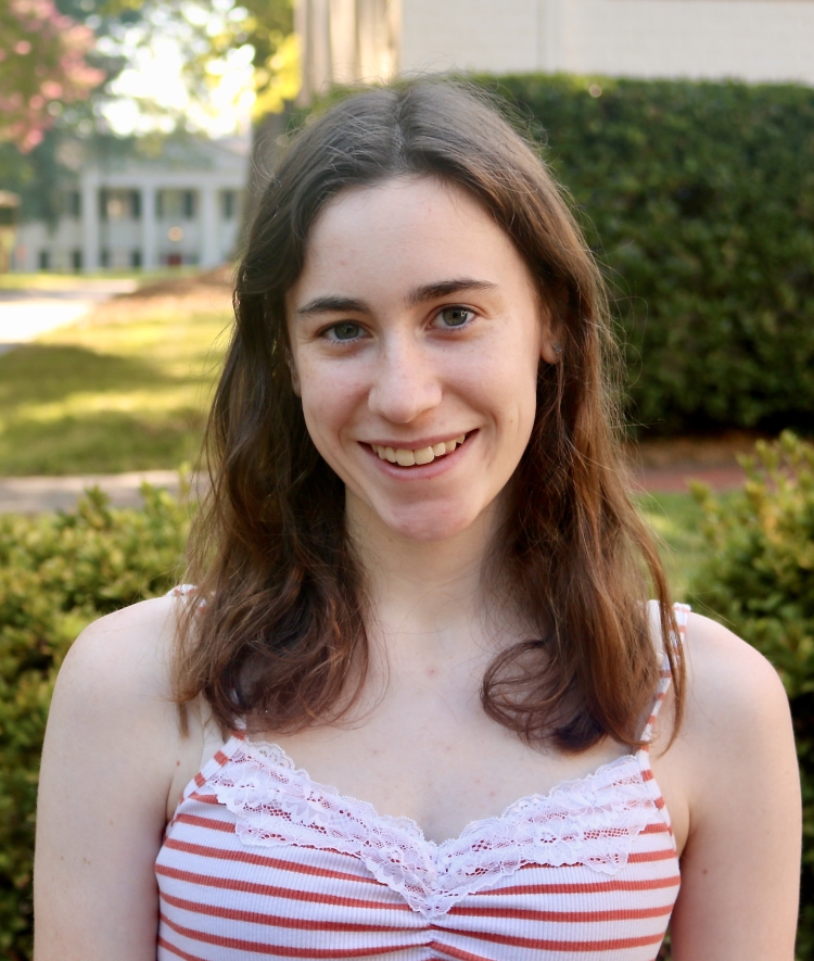 A smiling young woman with long, wavy brown hair, wearing an orange and white striped camisole with lace trim, stands outdoors in front of green bushes and a white, columned building.