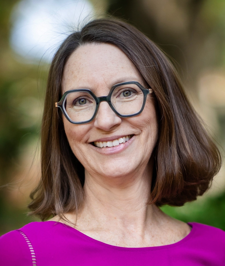 A woman with shoulder-length brown hair and glasses and a bright purple shirt