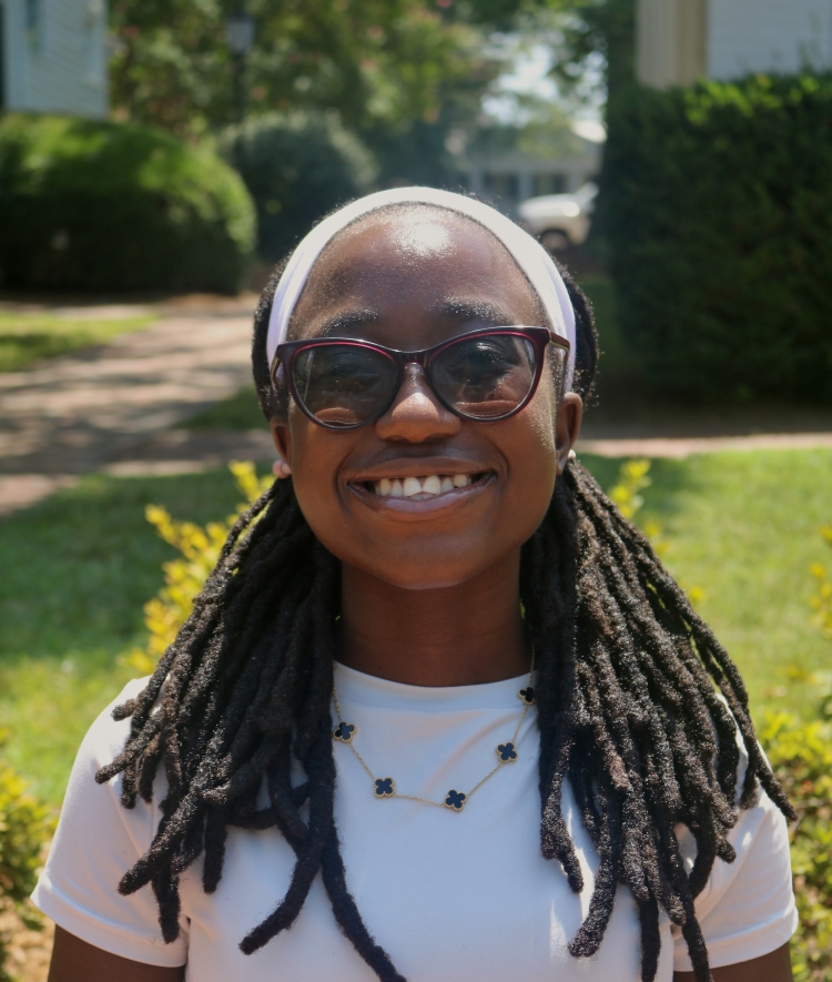 A smiling woman wearing dark sunglasses, a white headband over her dreadlocks, a white t-shirt, and a gold necklace with four-leaf clover pendants poses outside with blurred greenery and a path in the background.