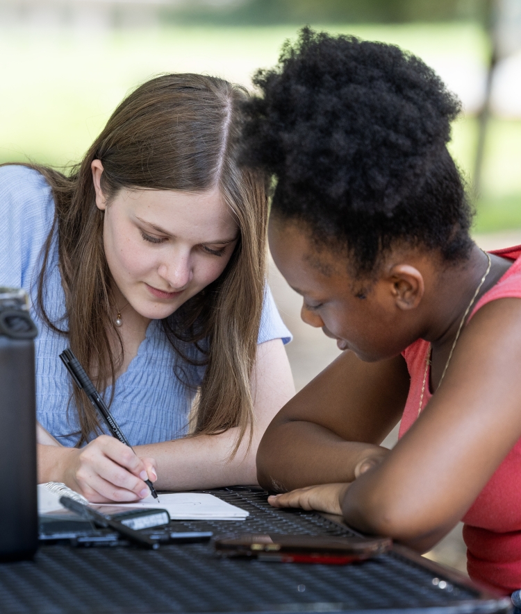 Two young women sitting at an outdoor table studying or writing together in a notebook.