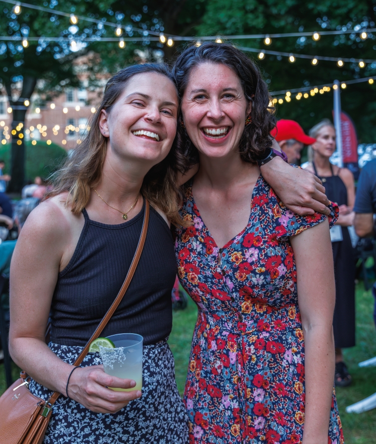 Two smiling women pose closely together at an outdoor evening event decorated with string lights overhead.