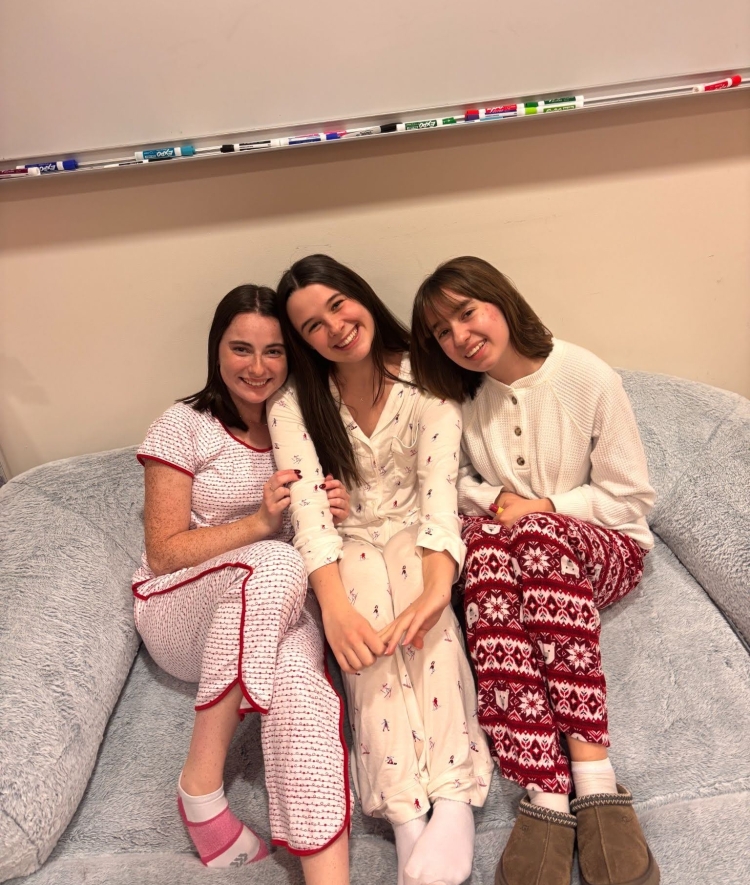 Three smiling young women sitting close together on a large, plush grey beanbag chair while wearing matching patterned pajamas.