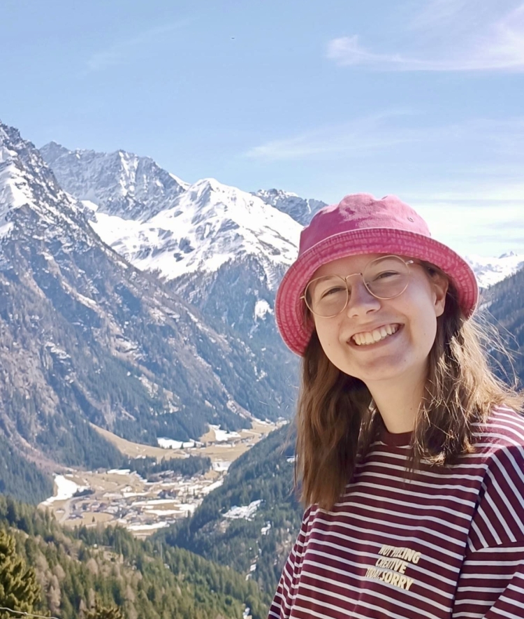A smiling person wearing glasses, a pink bucket hat, and a striped shirt stands in the foreground of a scenic mountain valley with snow-capped peaks in the background.