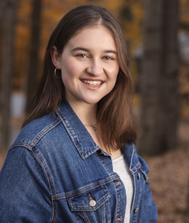 A smiling young woman with brown hair, wearing a denim jacket and hoop earrings, poses for a portrait against a blurred autumn background.