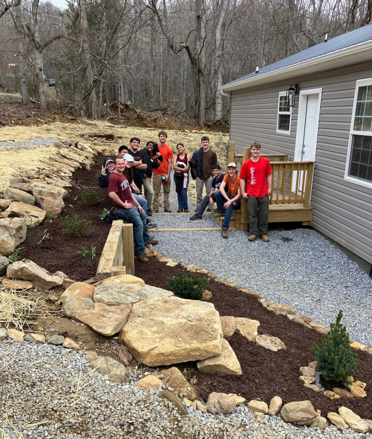 A group of volunteers stands along a newly constructed gravel pathway and stone retaining wall next to a house with light grey siding.
