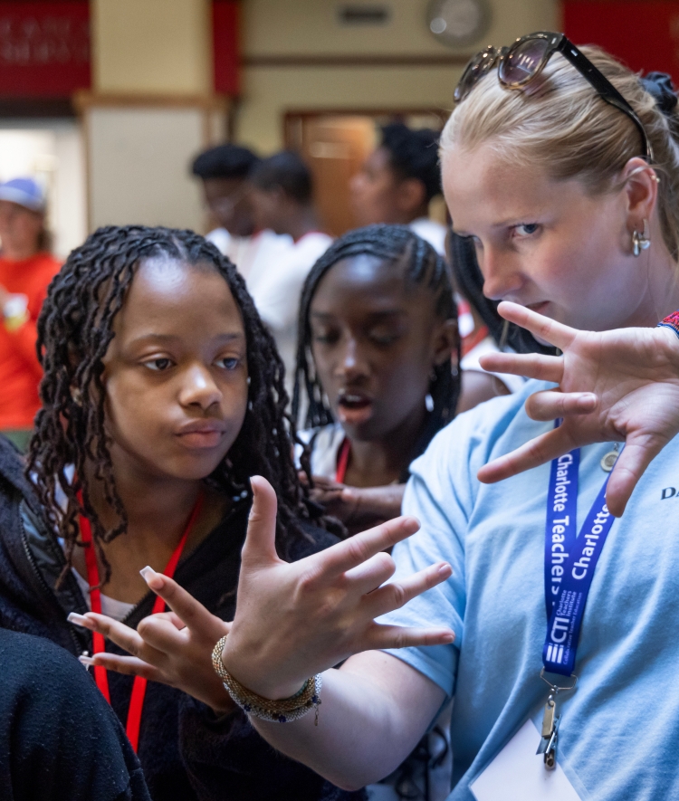 A instructor or staff member wearing a light blue Davidson College t-shirt and a Charlotte Teachers Institute lanyard demonstrates a hand sign (the ASL sign for "I love you" / sign language gesture) to two attentive young students.