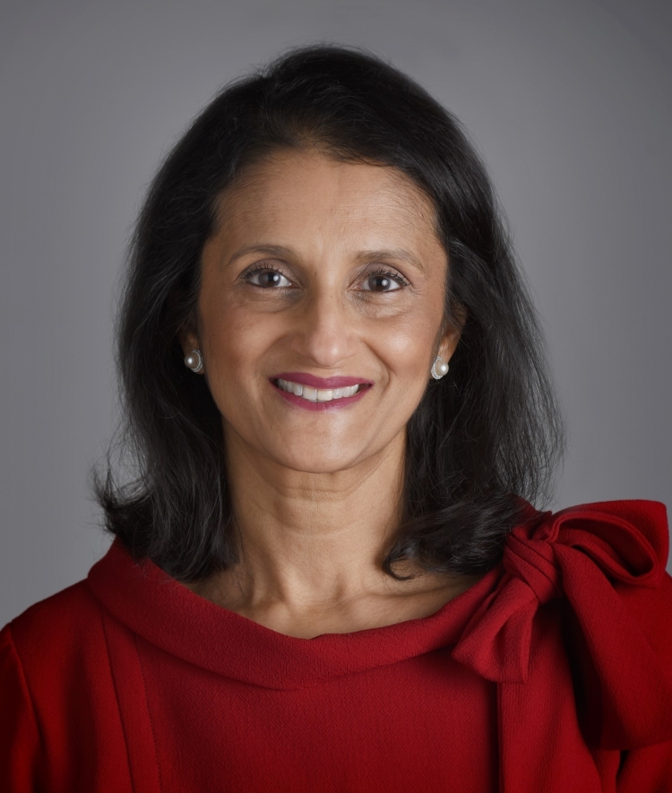 A portrait of Ameesha Pandya Kansupada smiling, wearing a red top with a bow on the shoulder against a neutral gray background.