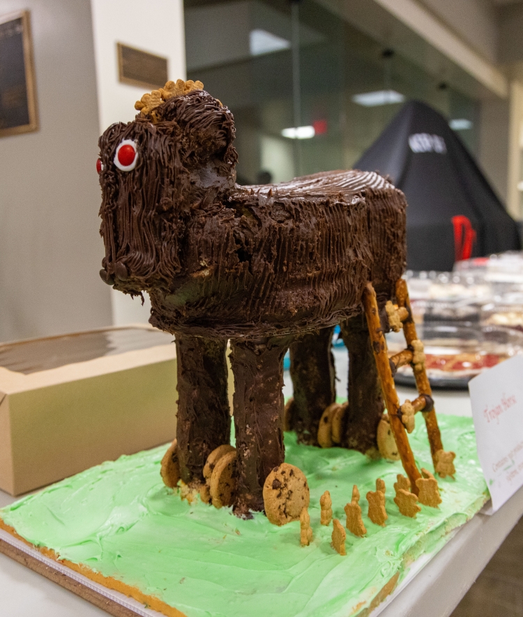 A chocolate-frosted cake shaped like a Trojan horse sits on a long display table alongside pretzel ladders, cookie wheels, and small cracker figures on green frosting.