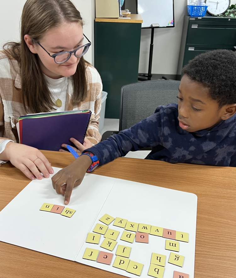 A student teacher sits beside a young student at a wooden desk as he points to letter tiles spelling "shin" on a folder.
