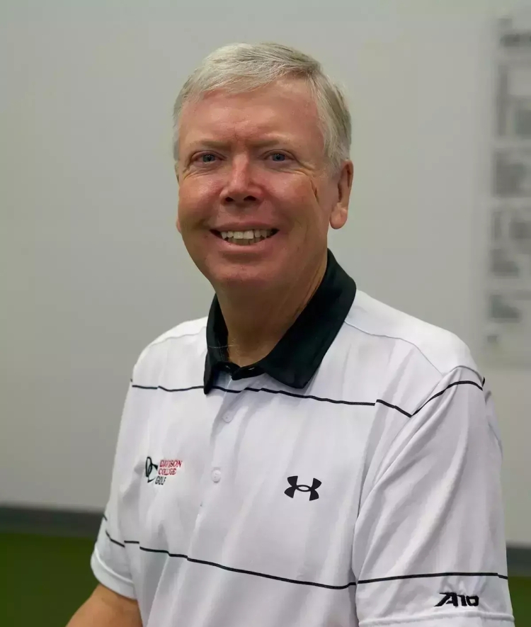 Portrait of a smiling man with short light-colored hair, wearing a white Under Armour polo shirt with thin dark horizontal stripes, a black collar, and Davidson College Golf and A10 logos.