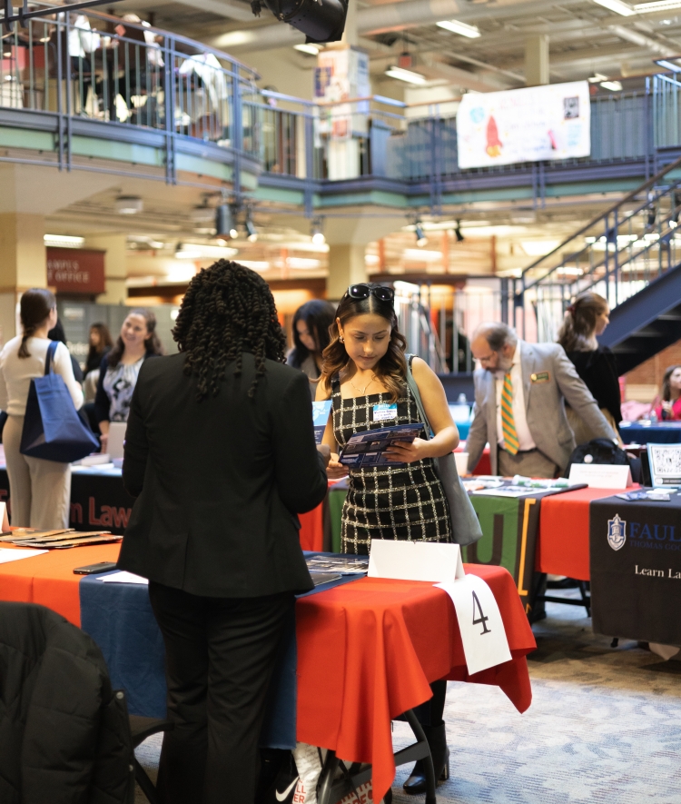 A woman holding promotional brochures speaks with a representative across a table numbered 4 at a multi-level law school fair.