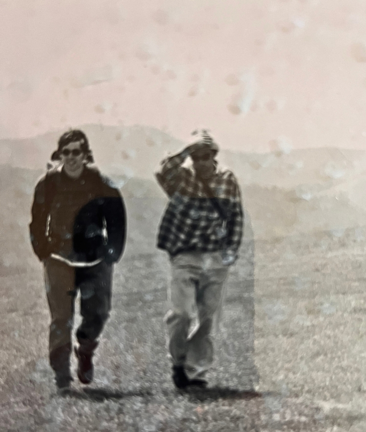 Two hikers walk through an open, grassy field on a mountain ridge with rolling hills in the background.