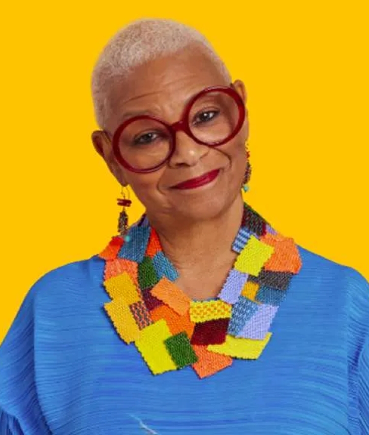 Portrait of North Carolina Poet Laureate Jaki Shelton Green wearing round red glasses, a colorful patch necklace, and a blue top against a bright yellow background.