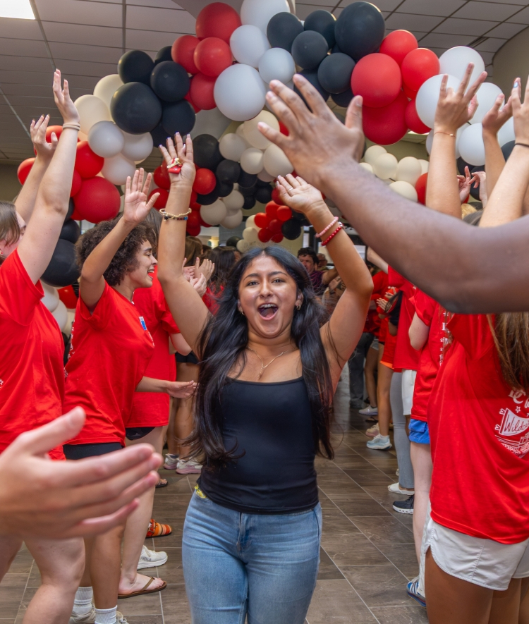 A young woman in a black tank top and jeans runs down a hallway filled with red, white, and black balloons while crowd members in matching red shirts cheer and high-five him.