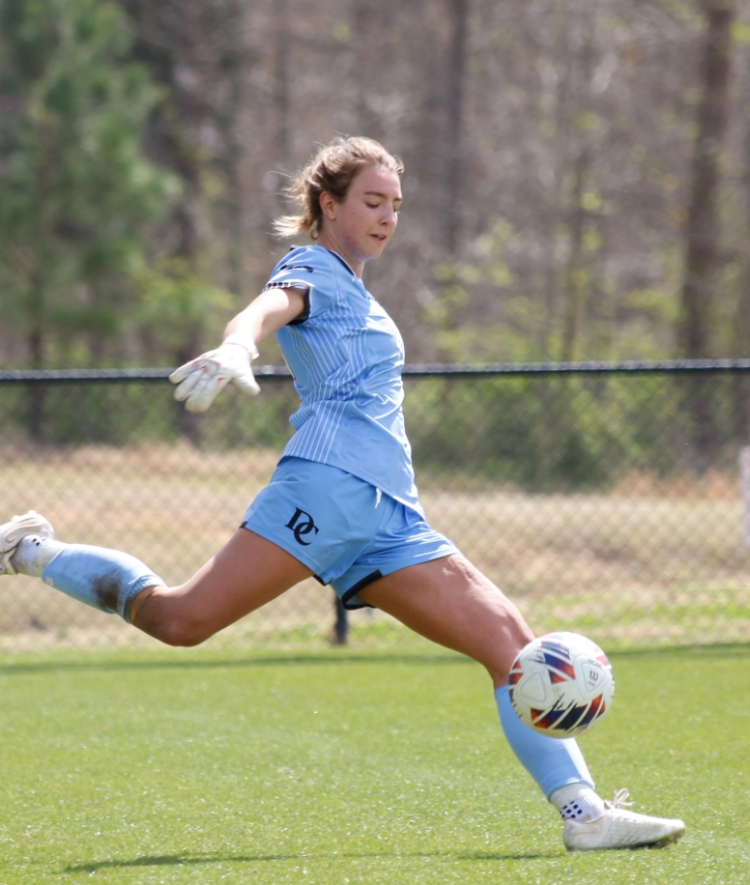 A female soccer goalkeeper in a light blue uniform kicks a soccer ball downfield on a grassy pitch during a sunny day.