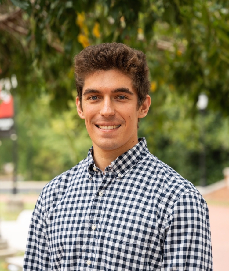 A young man with short brown hair and a warm smile stands outdoors wearing a blue and white checkered button-down shirt against a soft, blurred background of green foliage